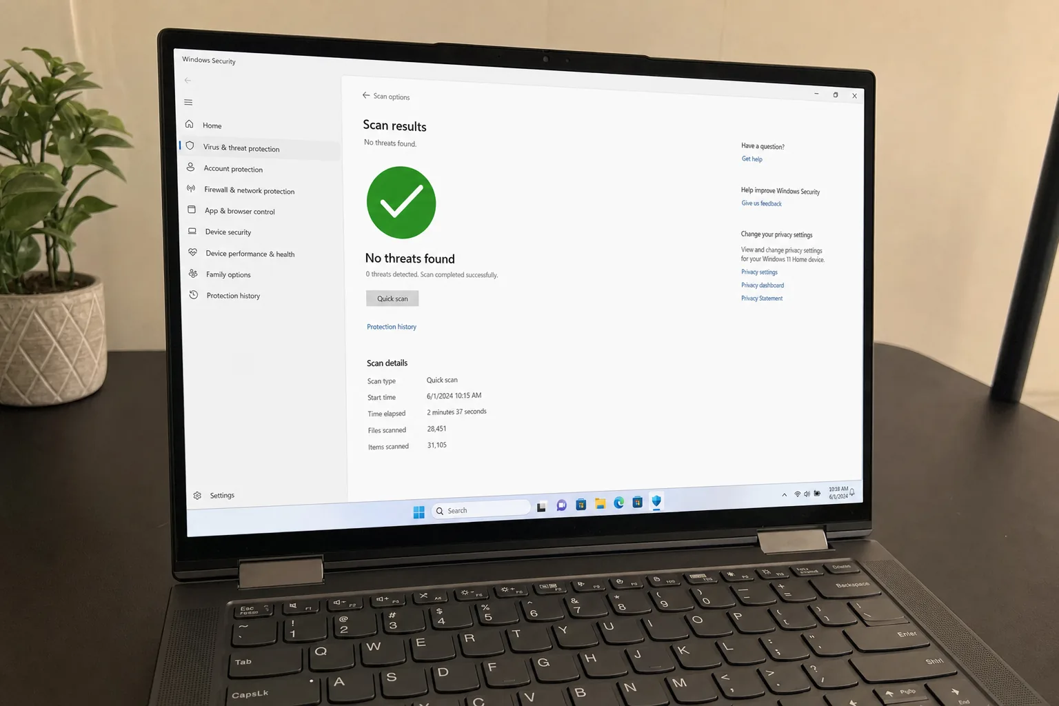 Laptop on dark desk showing Windows Security full scan complete with no threats found, snake plant in geometric planter beside it